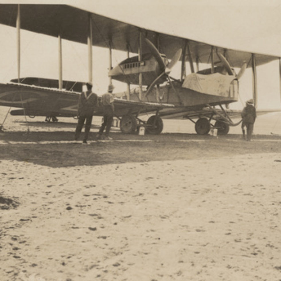 Image: Biplane on flat desert landscape. Tent off in the distance with armed guards.