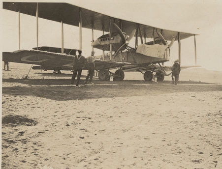 Image: Biplane on flat desert landscape. Tent off in the distance with armed guards.