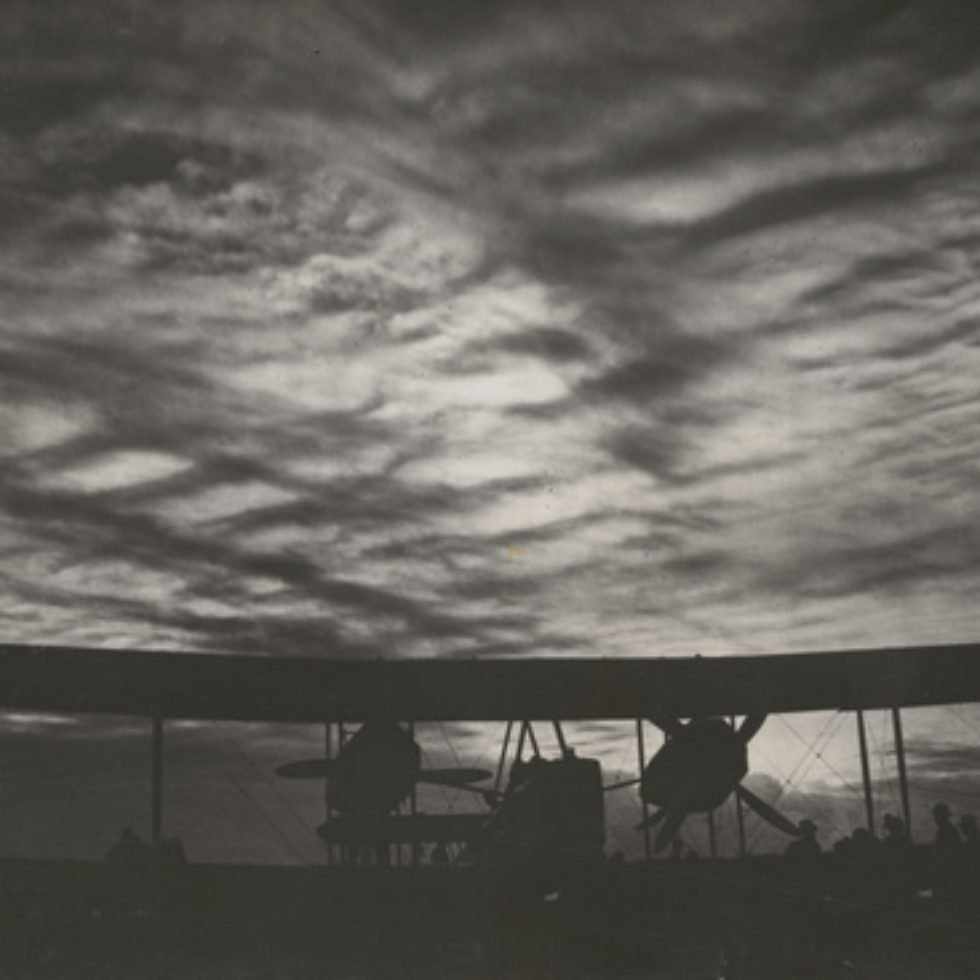 Image: Black and White photograph showing biplane with clouded sunset. Shadows of men gathered by the right wing