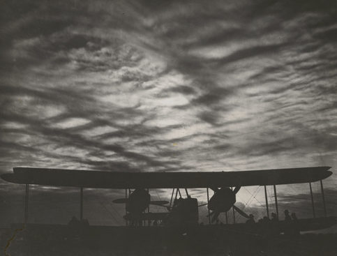 Image: Black and White photograph showing biplane with clouded sunset. Shadows of men gathered by the right wing